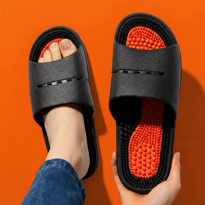 A foot wearing a black foot massage slipper next to one held by a hand.