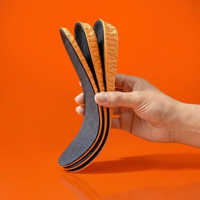 Height insoles in hand, featuring a gray top layer and orange edges, set against an orange background.